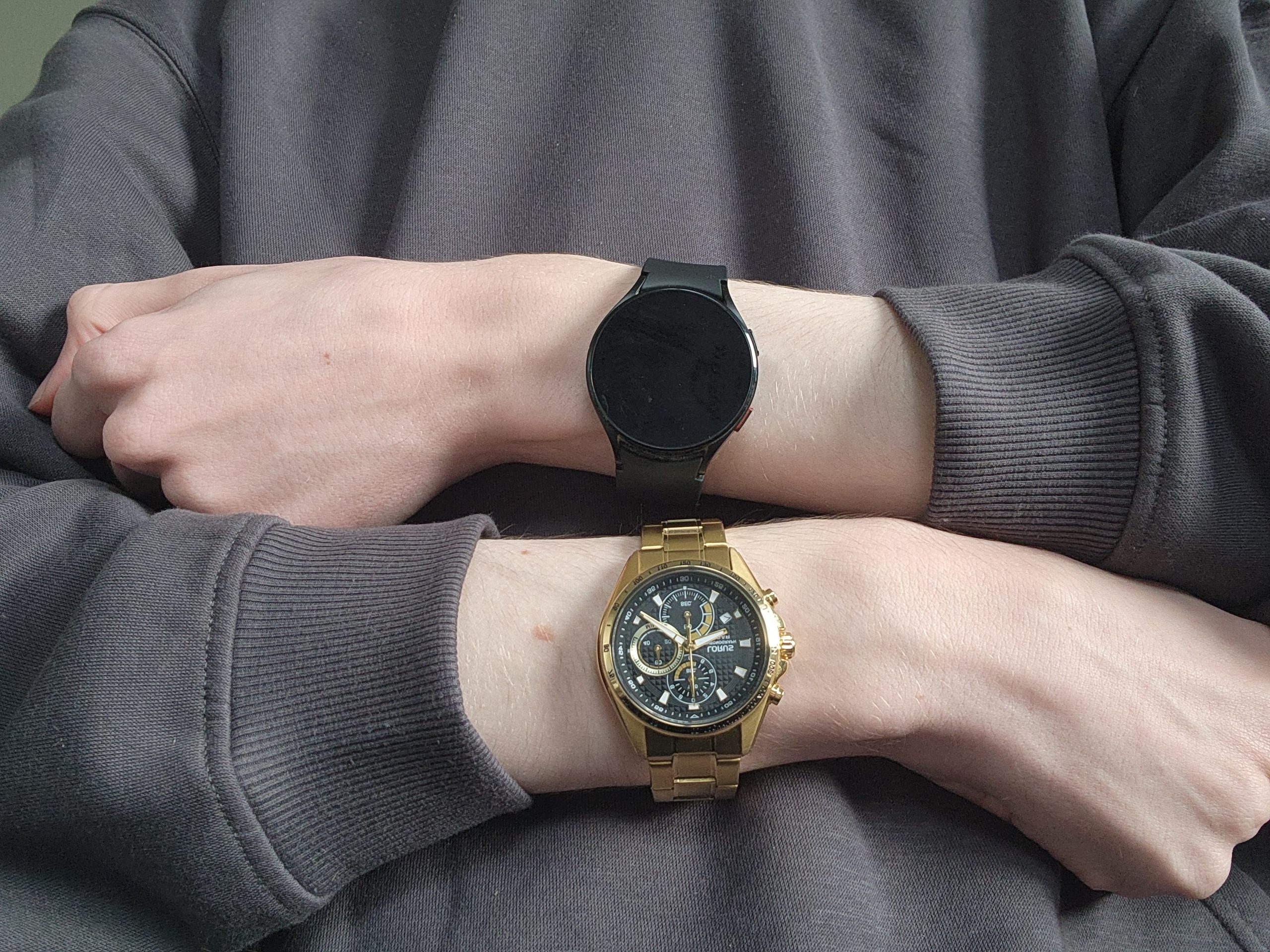 Braços cruzados sobre um moletom cinza, com um relógio em cada pulso: em cima um smartwatch preto de tela redonda apagada, embaixo um cronógrafo dourado de mostrador preto e pulseira de metal.