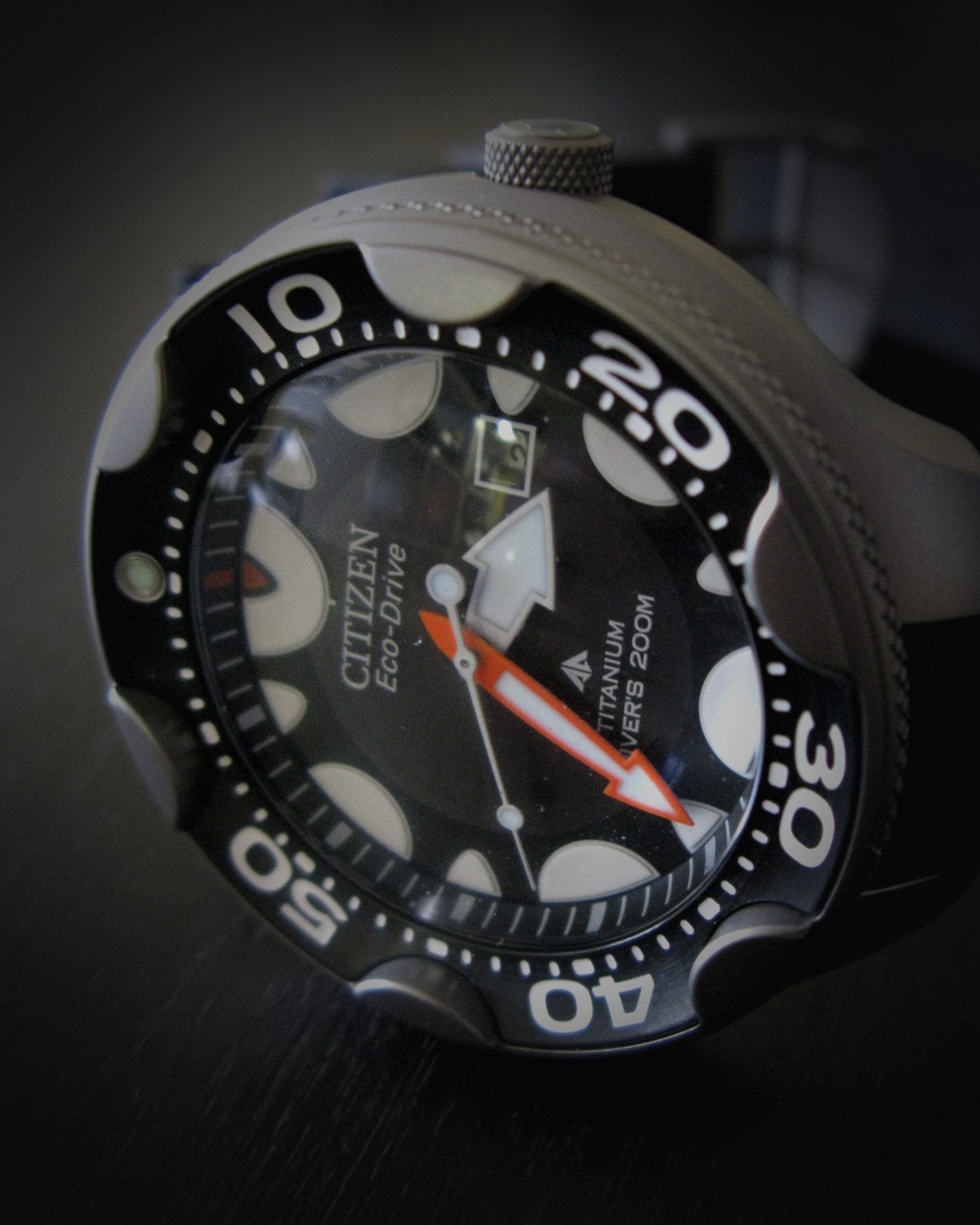 Arte da redação: relógio de mergulho Citizen Promaster Eco-Drive de titânio, enquadrado no mostrador, com a luz refeita e o fundo escurecido.