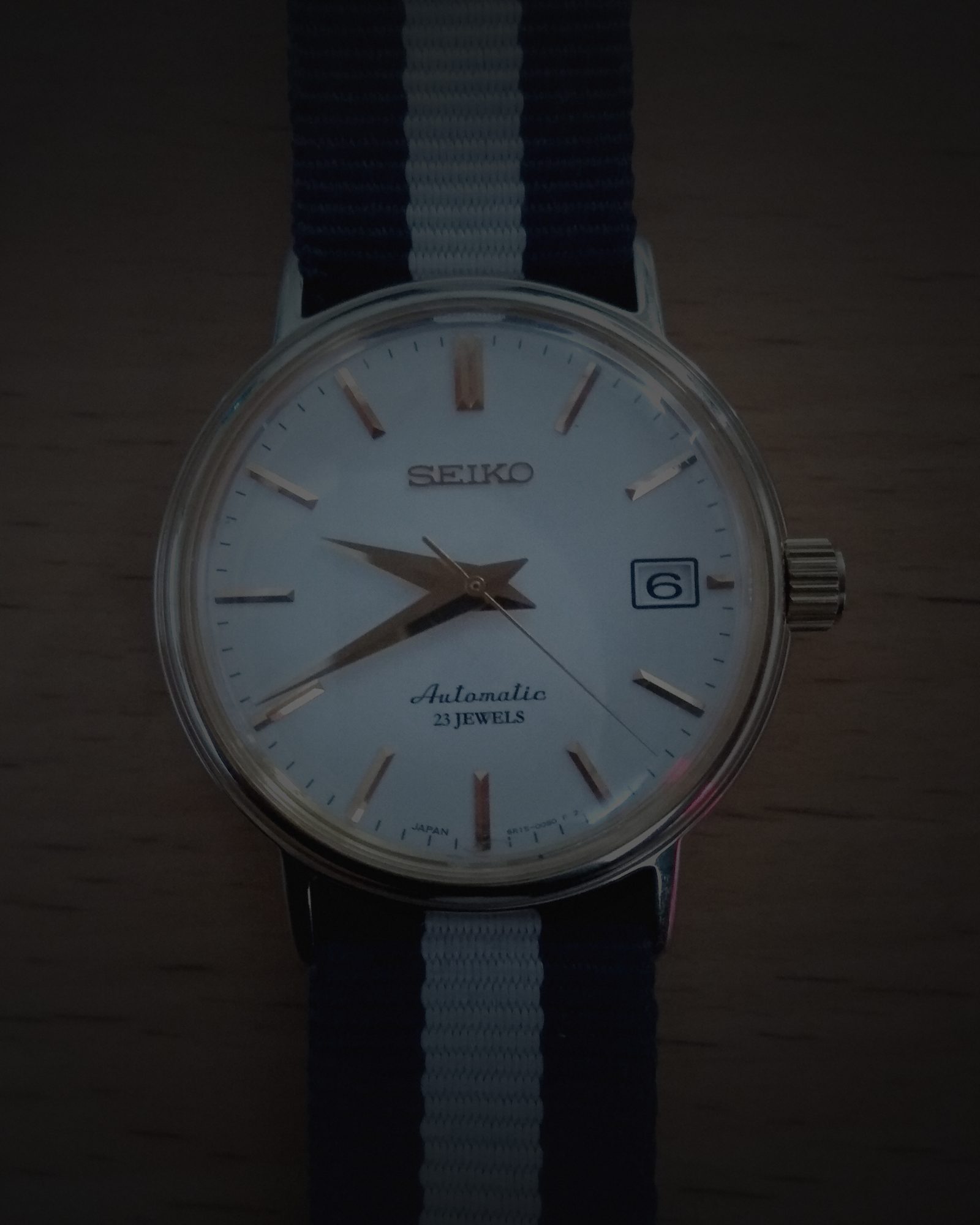 Arte da redação: relógio Seiko automático de mostrador branco e caixa dourada, enquadrado de perto sobre madeira, com a luz refeita e o fundo escurecido.