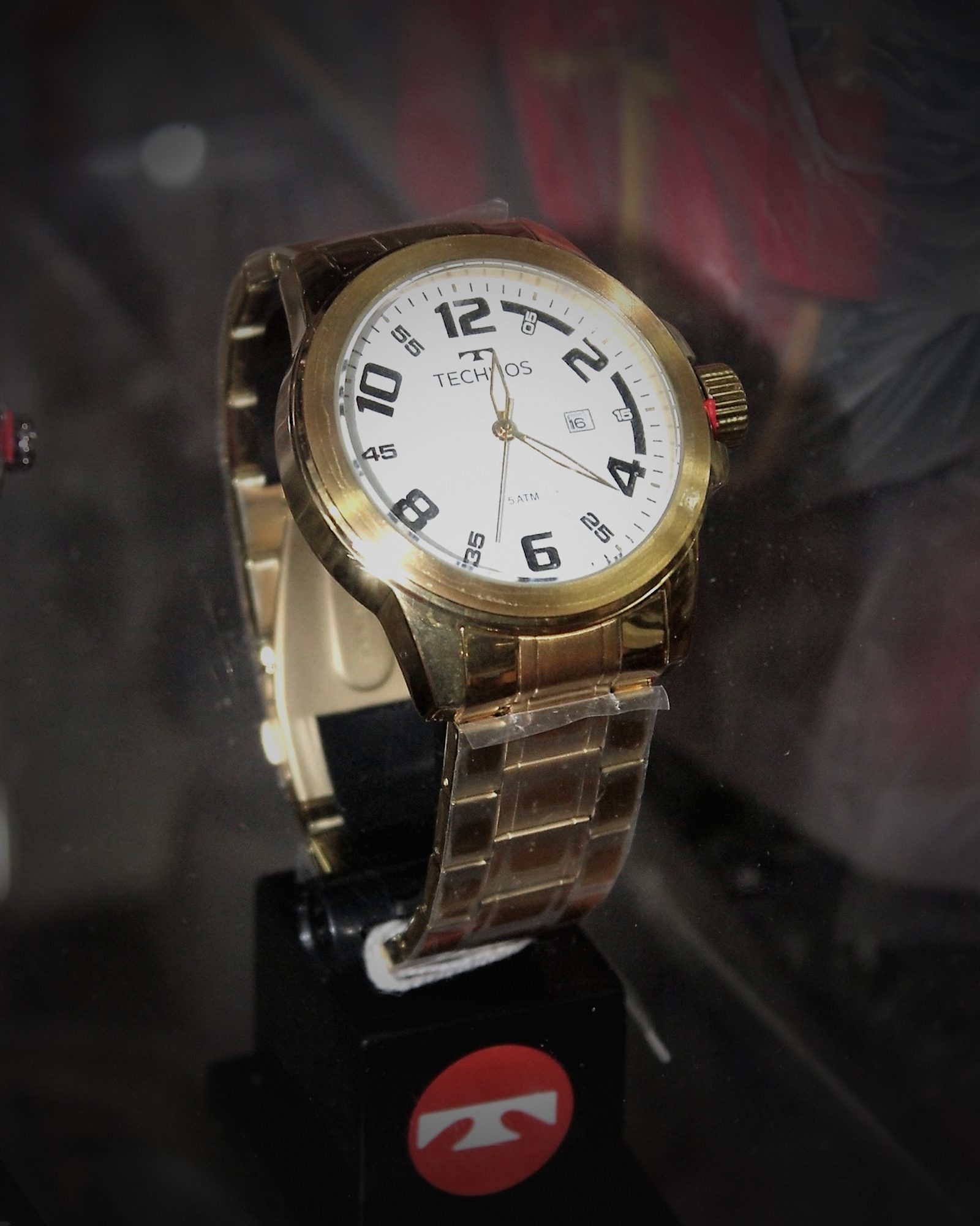 Arte da redação: relógio Technos dourado de mostrador branco, num expositor da marca, com a luz refeita e o fundo escurecido.